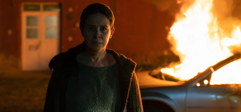 Eine Frau steht in der Nacht vor einem brennenden Auto und schaut entschlossen