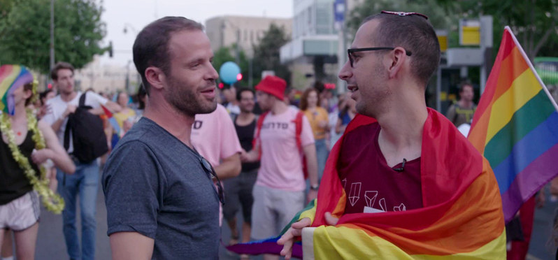 zwei Männer auf einer Pride-Demo unterhalten sich, einer trägt eine Regenbogenfahne und eine Kippa