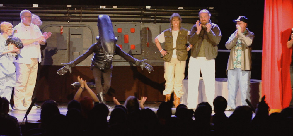 Bild Alien on Stage, ein als Alien verkleideter Schauspieler verbeugt sich auf der Bühne vor dem Publikum, daneben stehen weitere klatschende Darsteller:innen