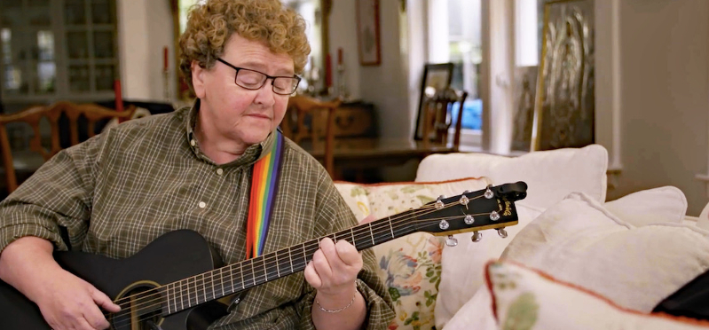 Eine Frau spielt auf einer Gitarre im Wohnzimmer, der Gurt der Gitarre ist in Regenbogenfarben