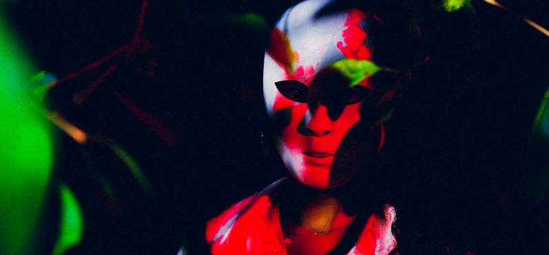 Eine maskierte Person ist halb versteckt hinter Pflanzen, sie wird von einem roten Licht beleuchtet