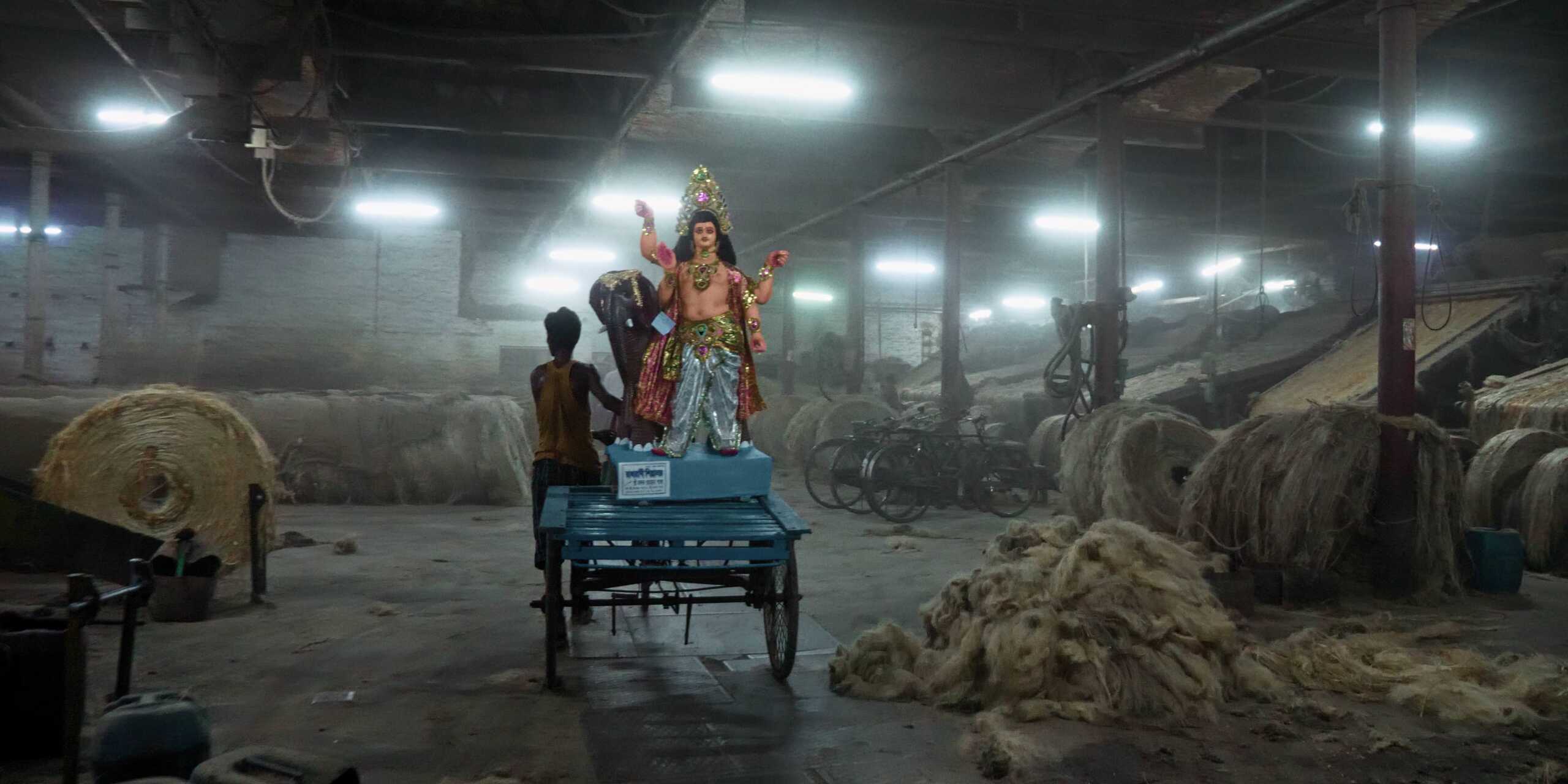 ein Arbeiter läuft neben seinem Gefährt in eine Jutefabrik hinein, auf dem Anhänger eine indische Göttinenfigur, große Juteballen an den Seiten
