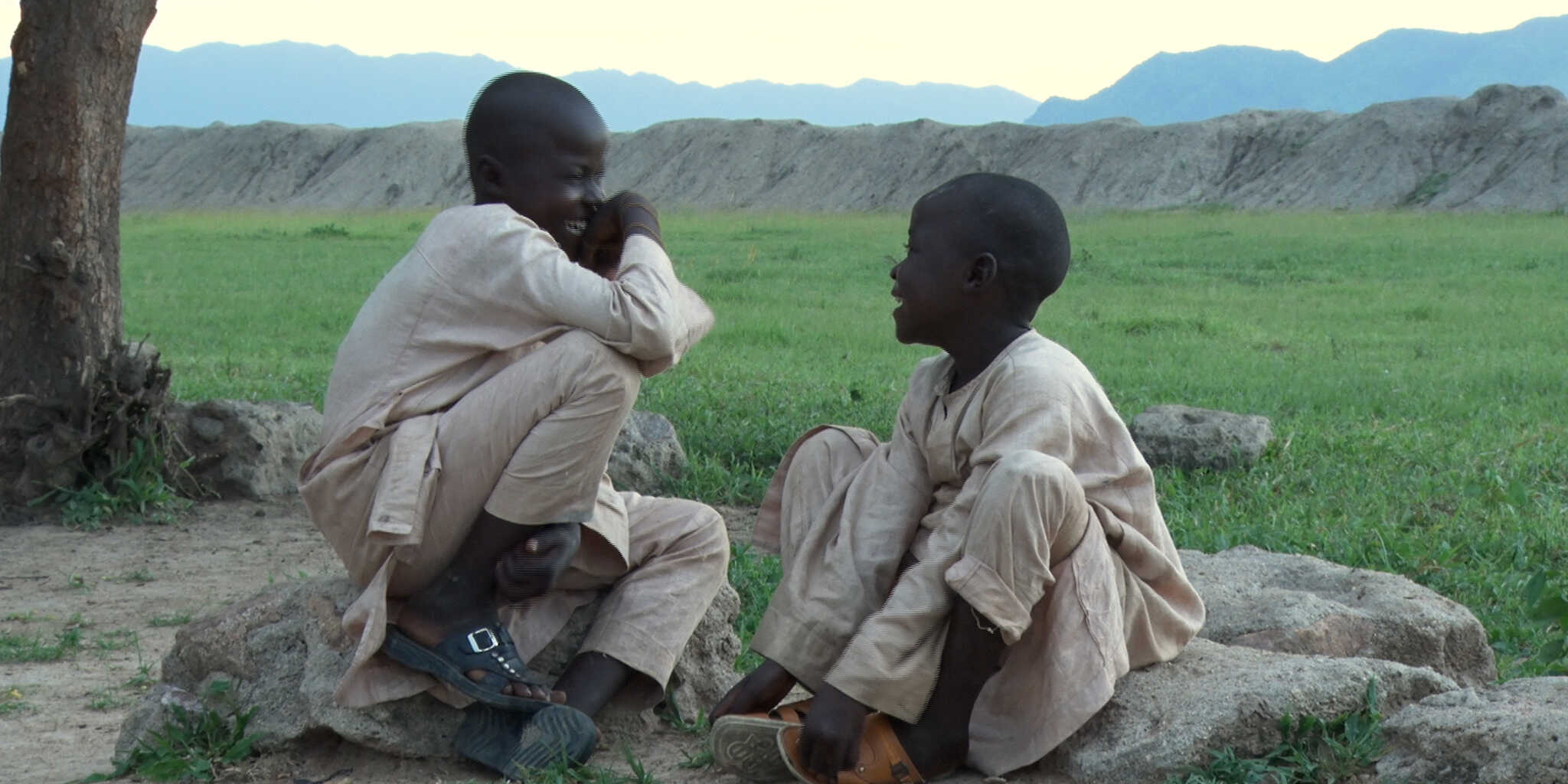 Zwei Jungen sitzen sich auf ein paar Steinen vor einer großen Wiese gegenüber und lachen