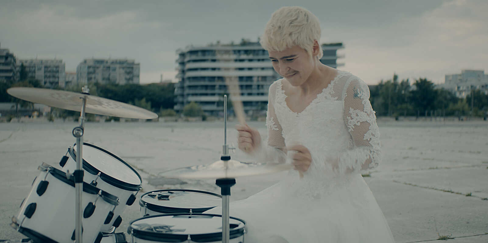 Eine Musikerin im Brautkleid spielt Schlagzeug auf einem weiten Platz, im Hintergrund Hochhäuser