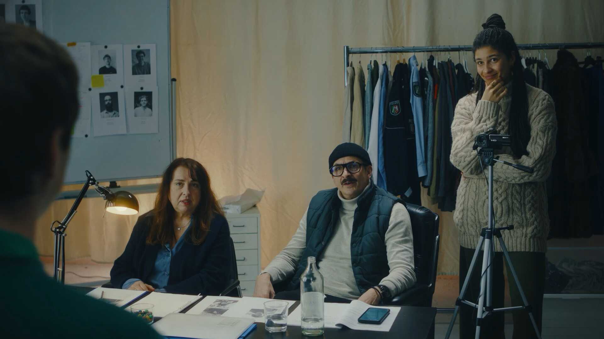 Casting: Mann und Frau sitzen am Tisch, eine Frau steht hinter einer Kamera. Im Hintergrund Setkarten und Kostüme.